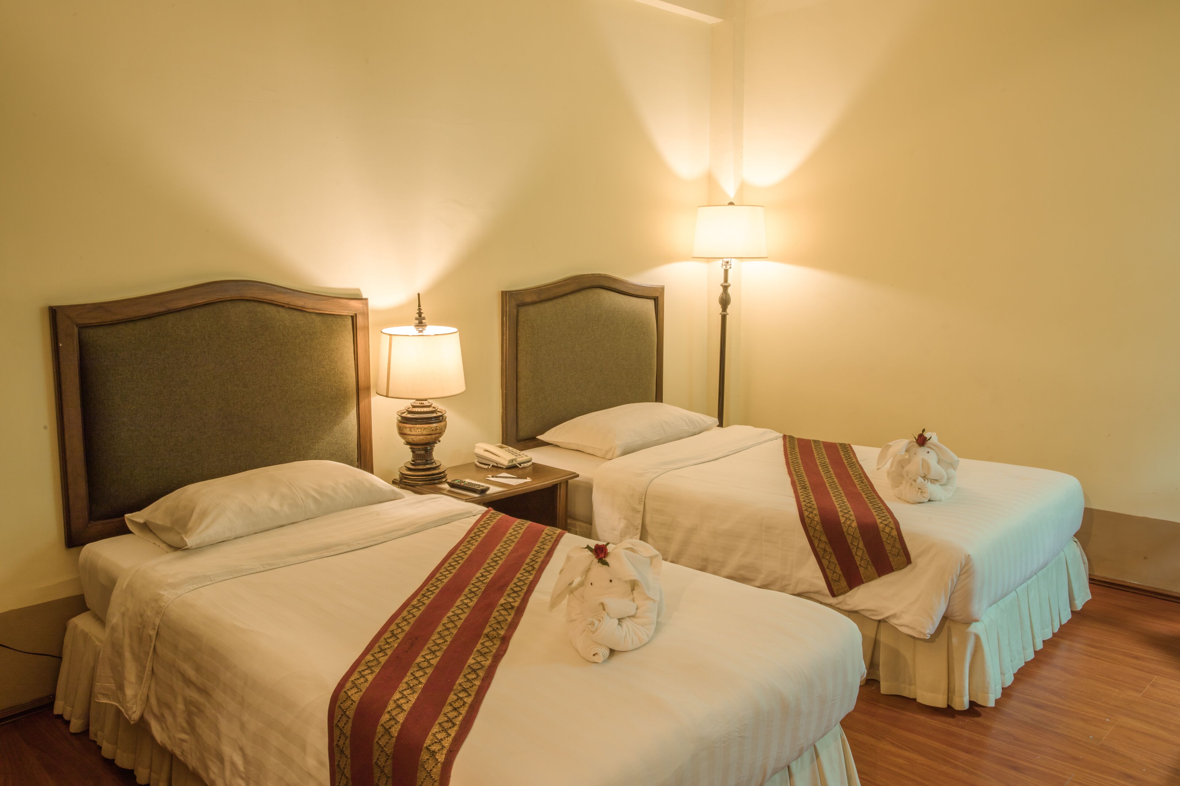 A hotel room with two beds, each with a headboard, bedside tables, lamps, and a decorative towel with a flower.
