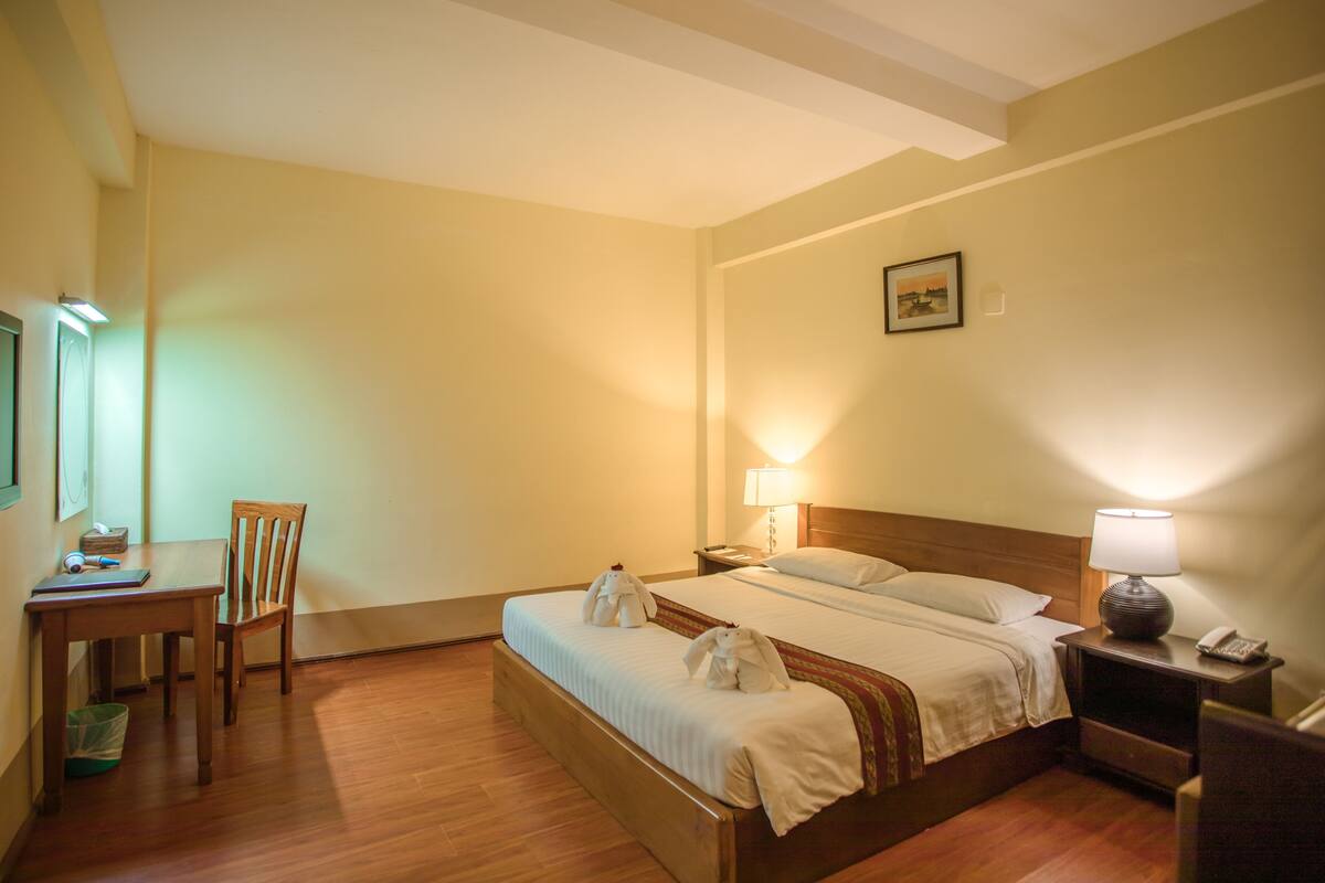 A hotel room with a bed, bedside lamps, a desk with a chair, a television, and a framed picture on the wall.