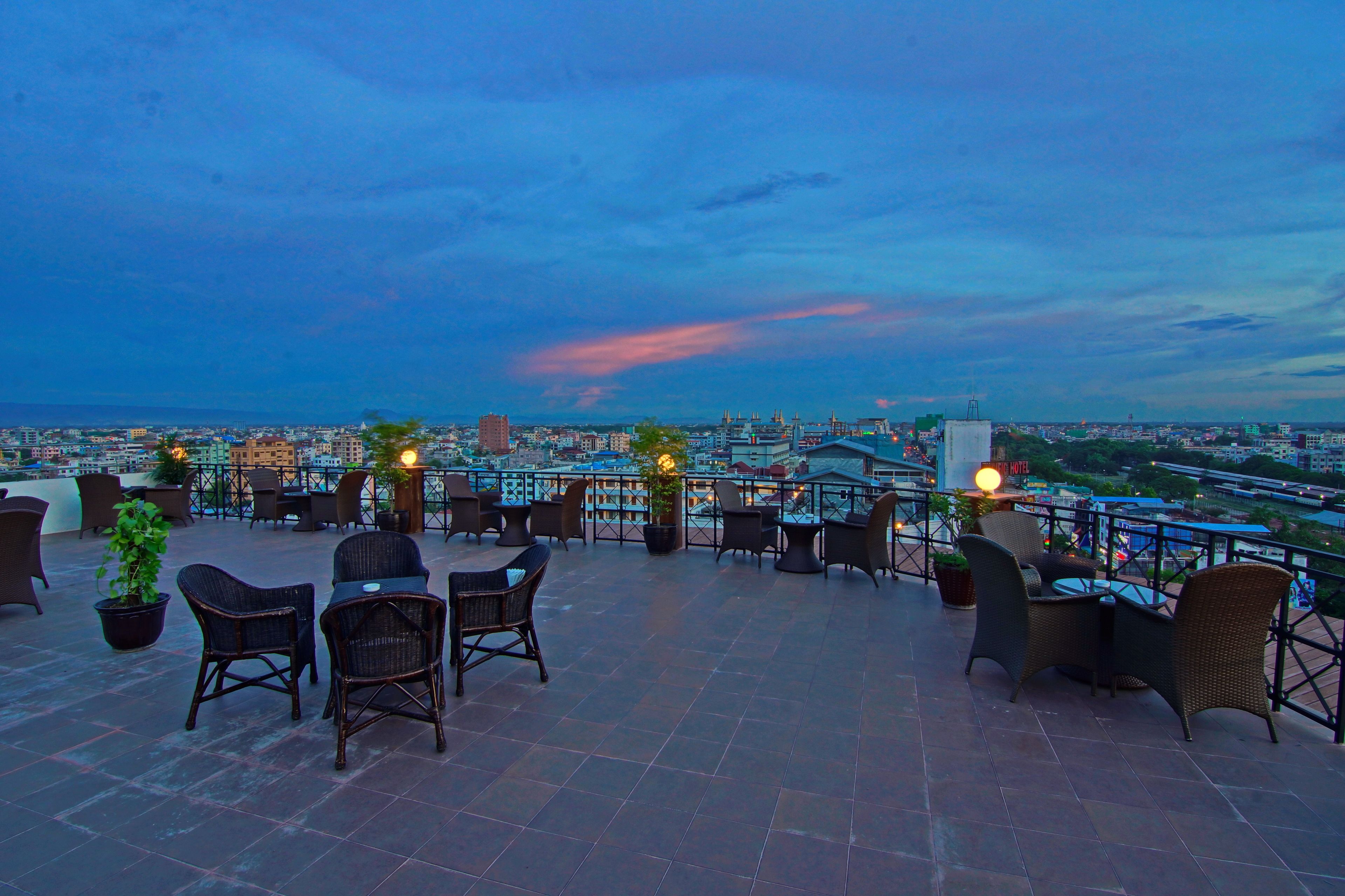 A rooftop terrace with wicker furniture and potted plants, overlooking a cityscape at dusk.