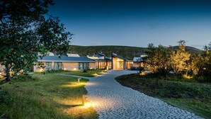 Ett modernt hus med en asfalterad uppfart som leder till ett garage, omgivet av grönska och kullar i bakgrunden.