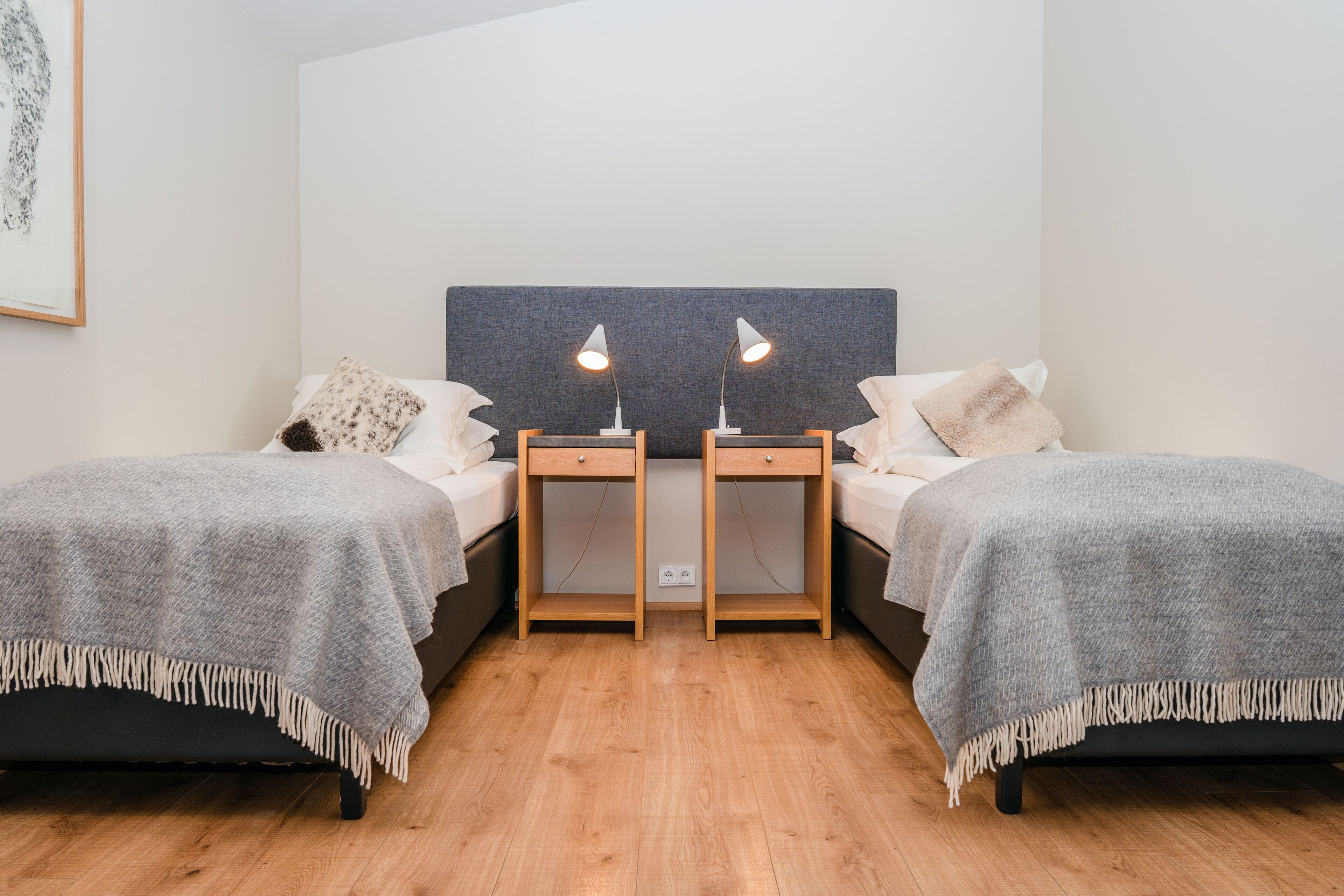 A double room with two beds, each with a bedside table and lamp, a grey headboard, and a wooden floor.