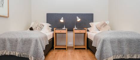 A double room with two beds, each with a bedside table and lamp, a grey headboard, and a wooden floor.