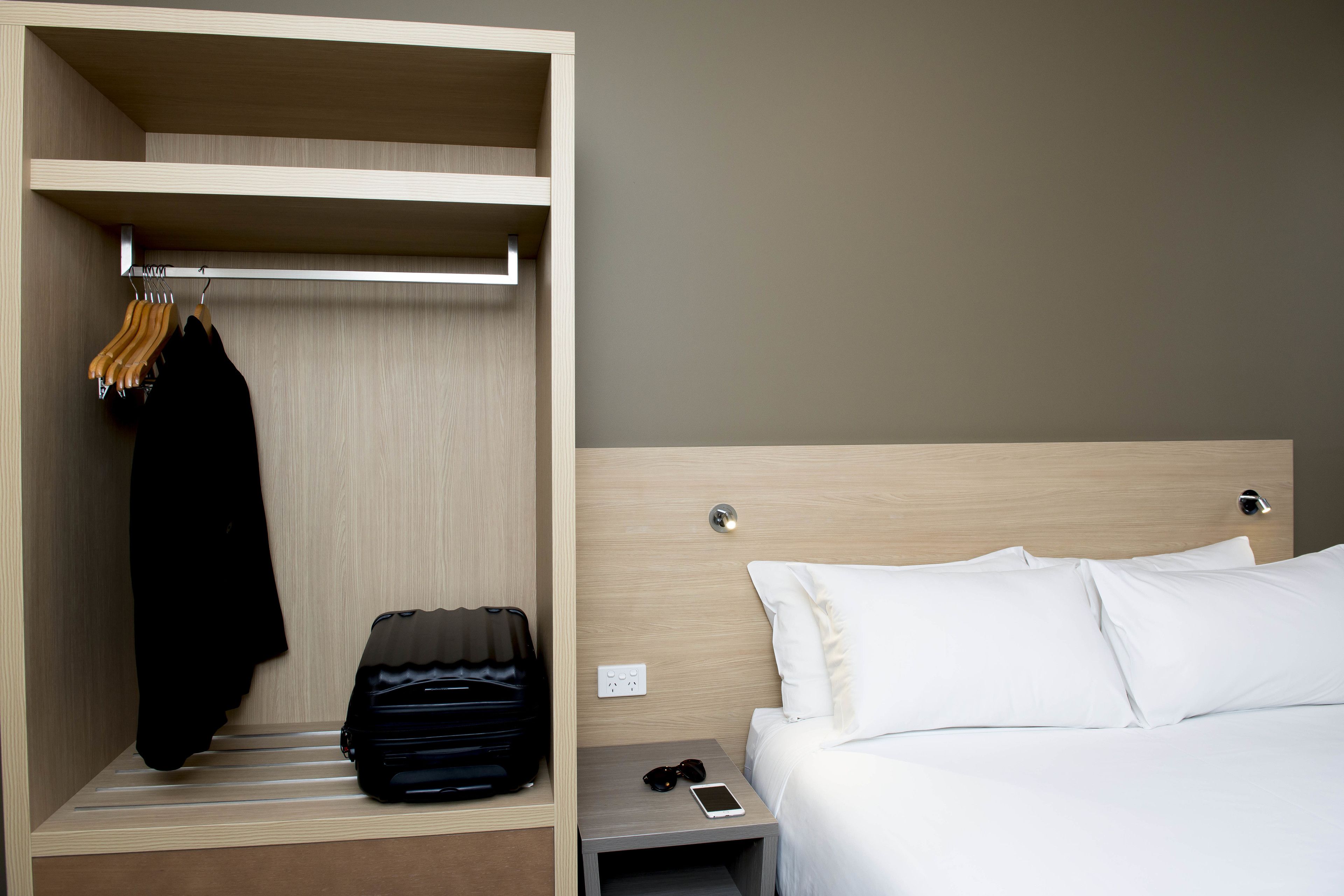 Una habitación de hotel con un armario de madera, una cama con ropa blanca, una mesita de noche con un teléfono y gafas de sol, y una maleta en el suelo.