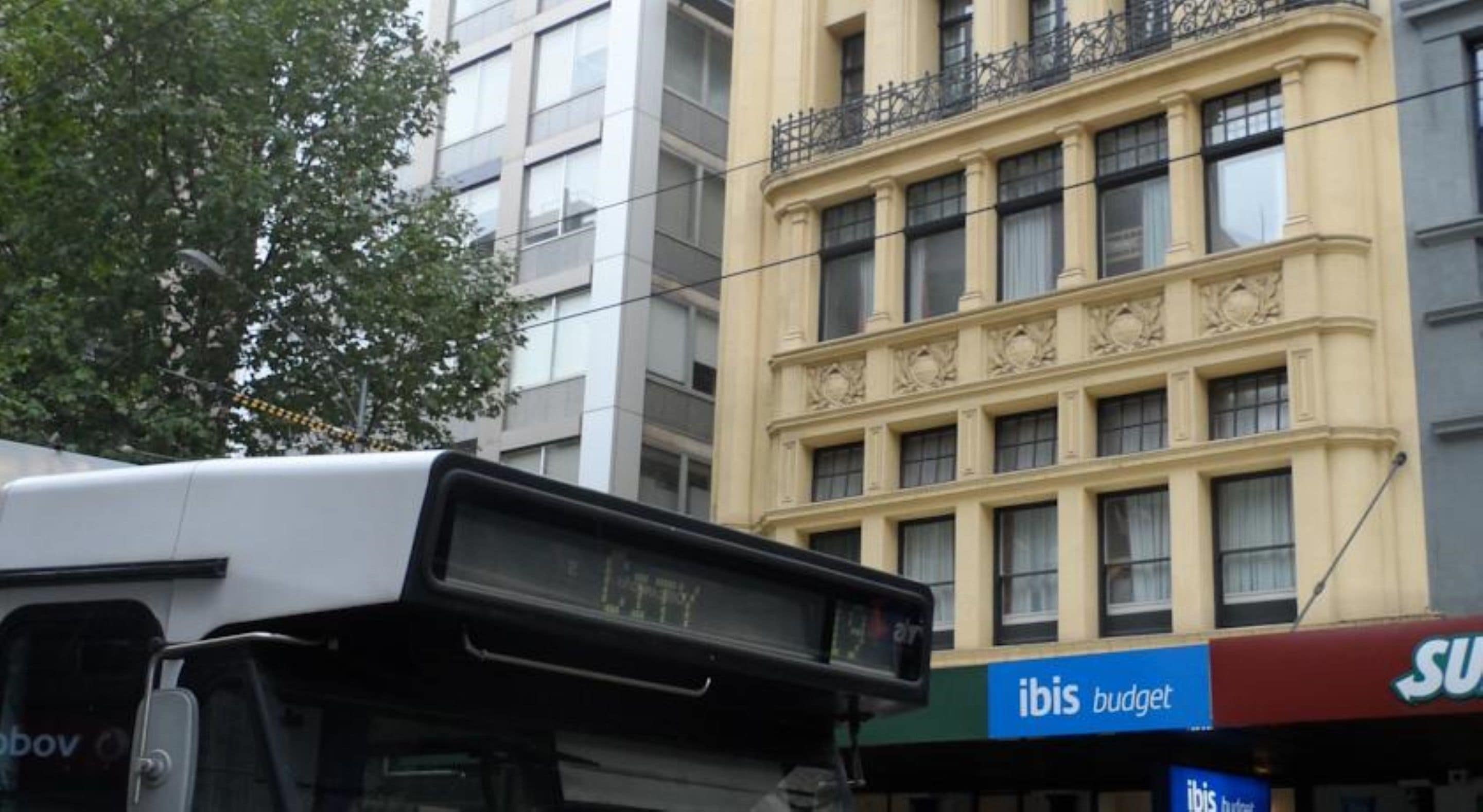 Un autobús amb pantalla digital, un edifici amb el rètol 'ibis budget' i un arbre al fons.