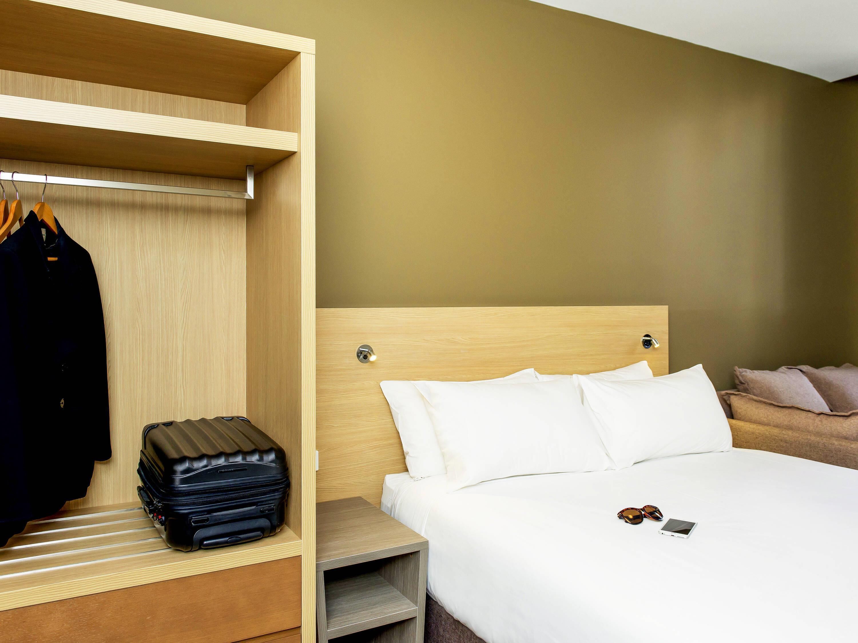 A hotel room with a bed, a wooden wardrobe, and a small bedside table.