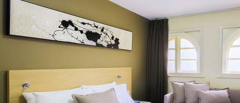 A hotel room with a large bed, a smaller bed, and a sofa. There is a painting on the wall and a window with curtains.