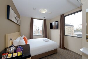 Quarto de hotel com cama, televisão de tela plana, janela com cortinas, cafeteira e uma pequena mesa com objetos.