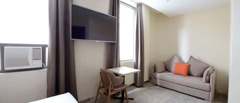 A hotel room with a bed, a desk, a chair, a sofa, a flat-screen TV, and a window with curtains.