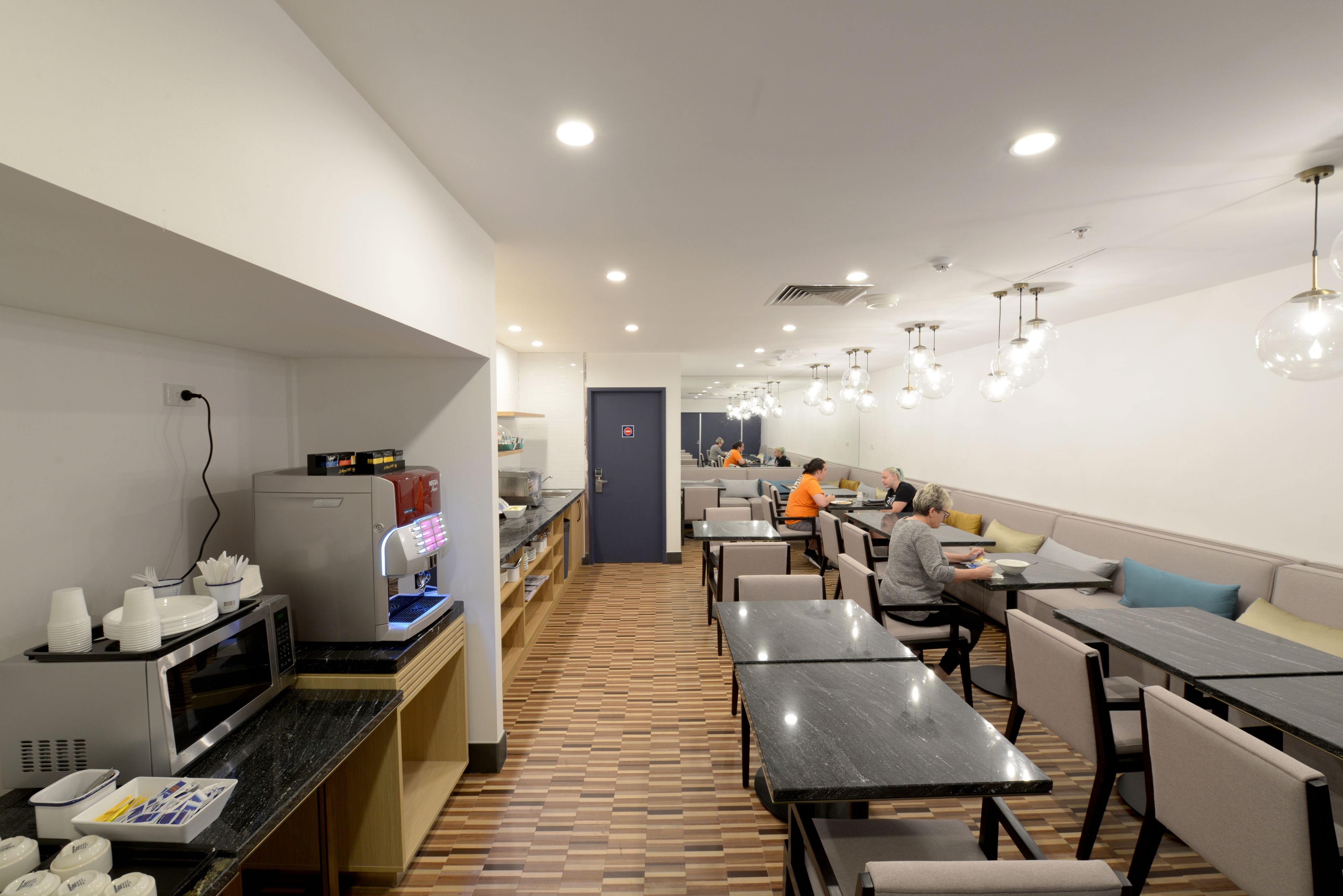 Een modern restaurant met lange gedeelde tafels en zitjes, een koffiemachine en een toonbank met servies.