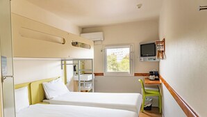 Une chambre d’hôtel compacte comprenant deux lits, un petit bureau avec une chaise, une télévision et une fenêtre laissant entrer la lumière naturelle.