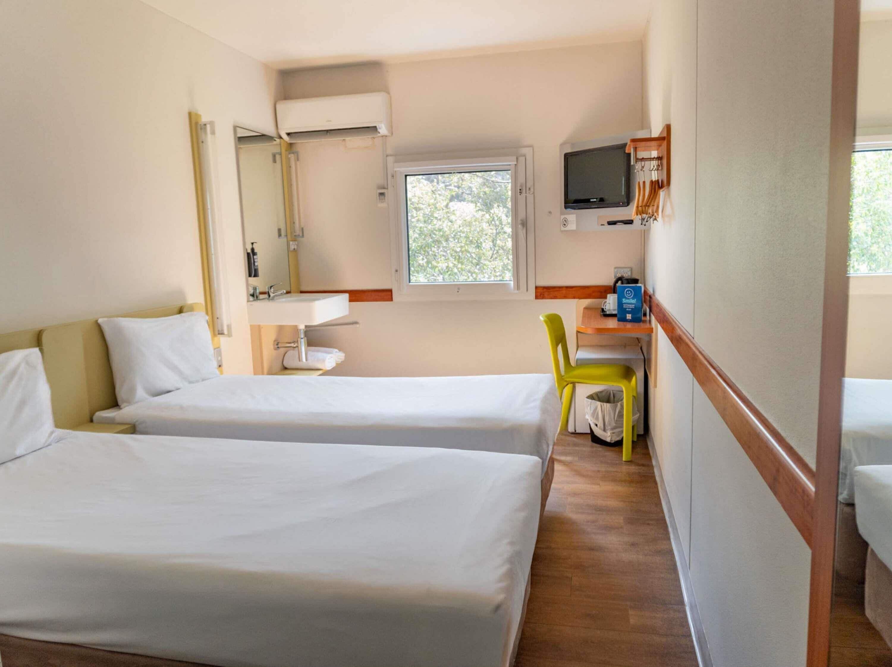 A hotel room with two beds, a bathroom with a mirror and sink, a TV mounted on the wall, and a window providing natural light.