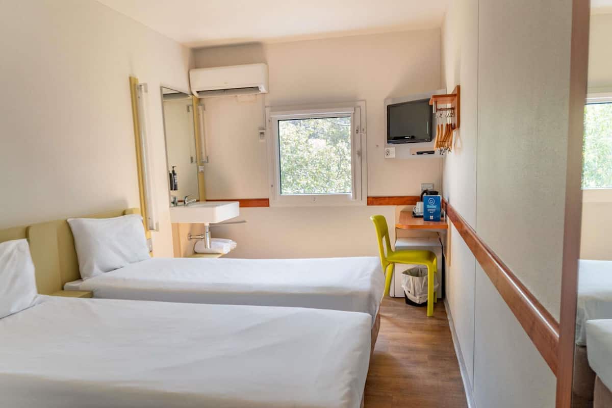 Une chambre d’hôtel avec deux lits, une salle de bain avec un miroir et un lavabo, un téléviseur fixé au mur et une fenêtre laissant entrer la lumière naturelle.