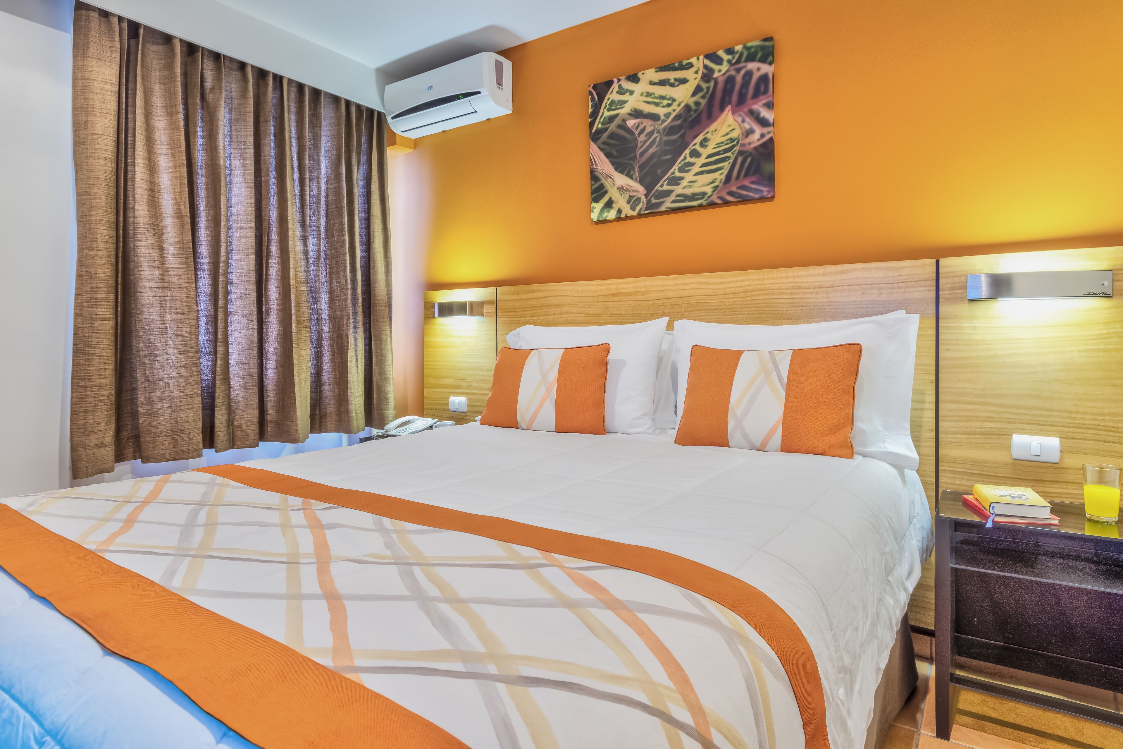 A hotel room with a large bed, an air conditioning unit, a nightstand with a glass of orange juice, and a painting on the wall.