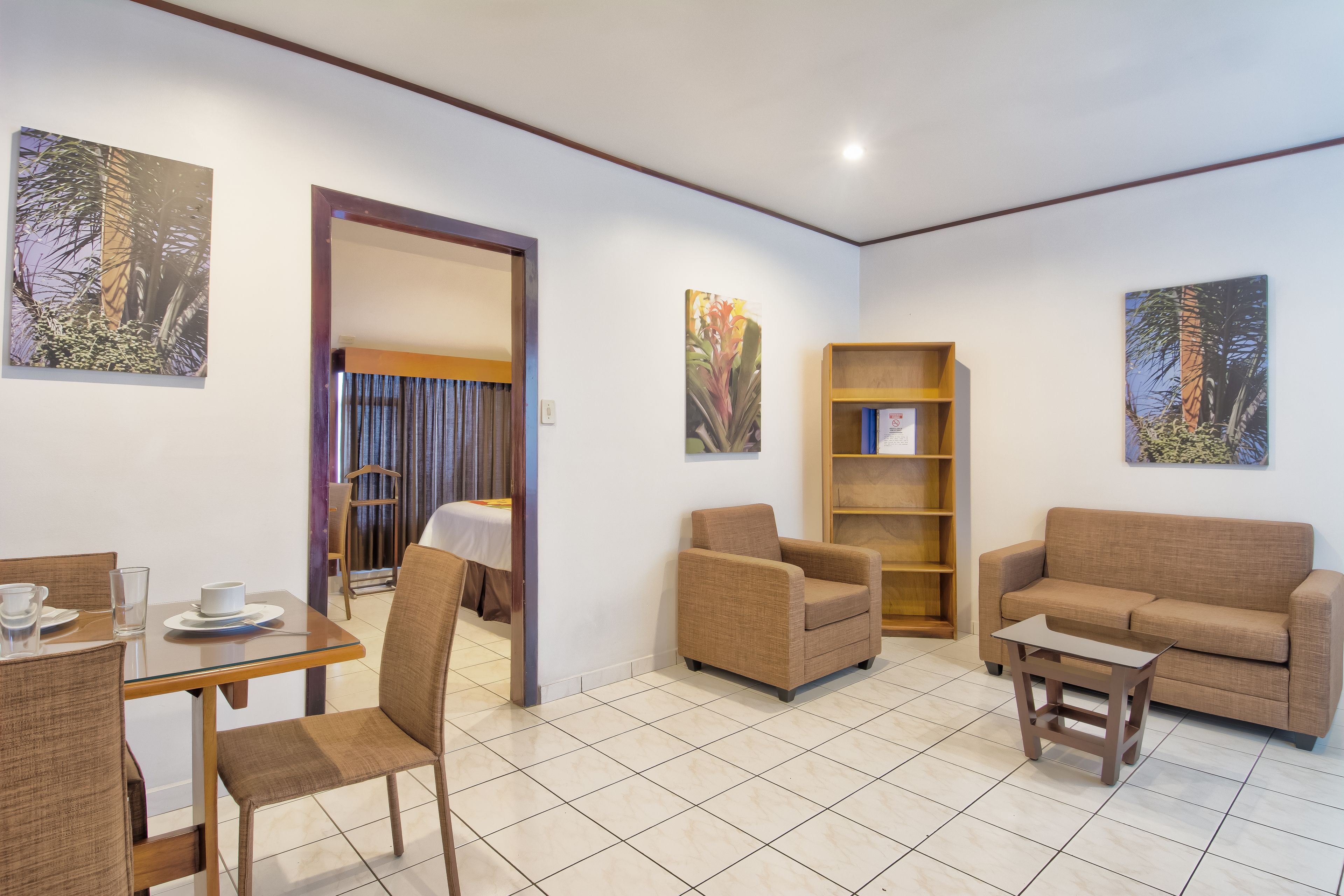 A furnished living room with a sofa, armchair, and a small table. There is a bookshelf and a dining area with chairs and a table.