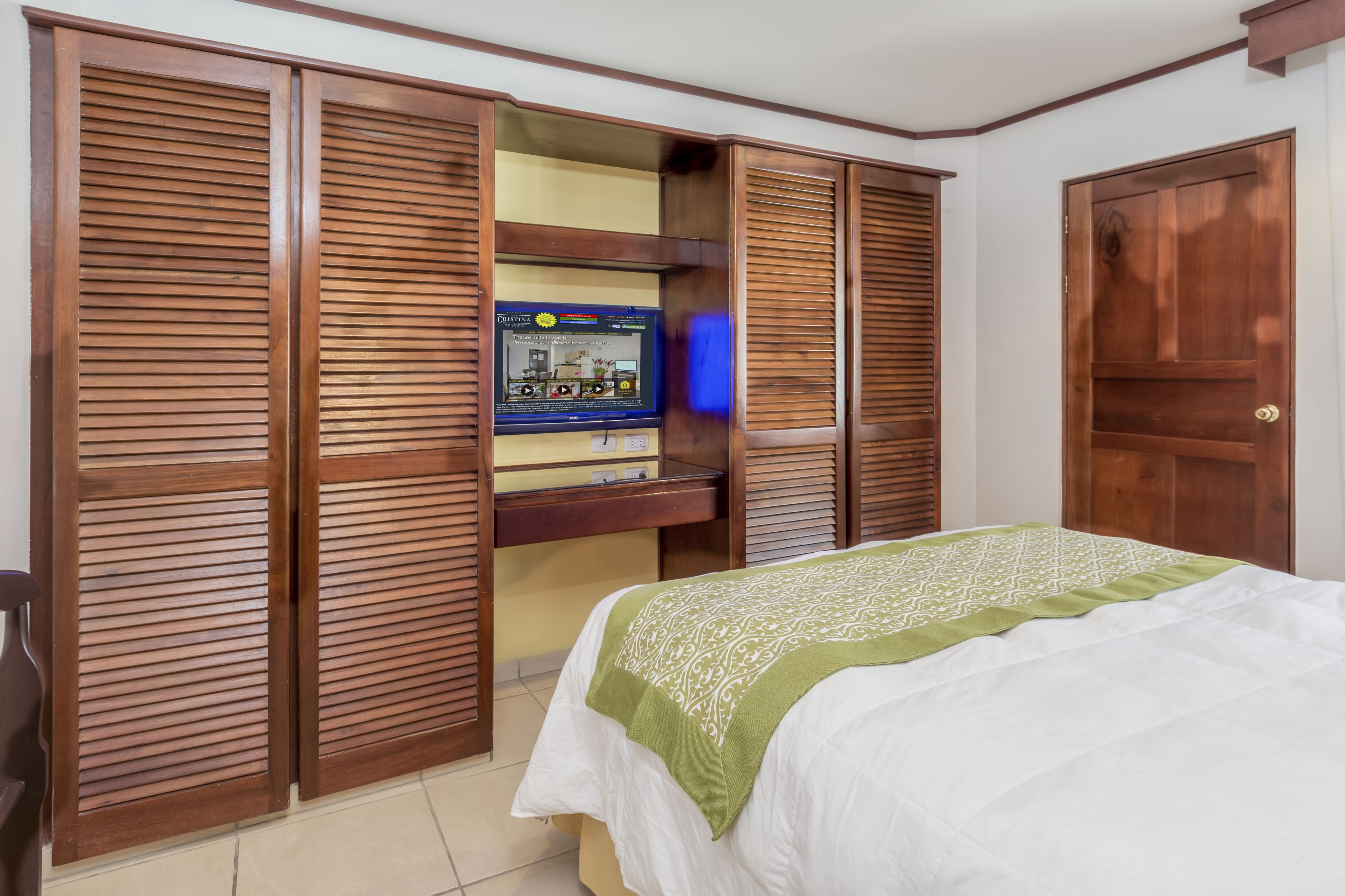 A bedroom with a bed, wooden wardrobe with louvered doors, and a television on a shelf.