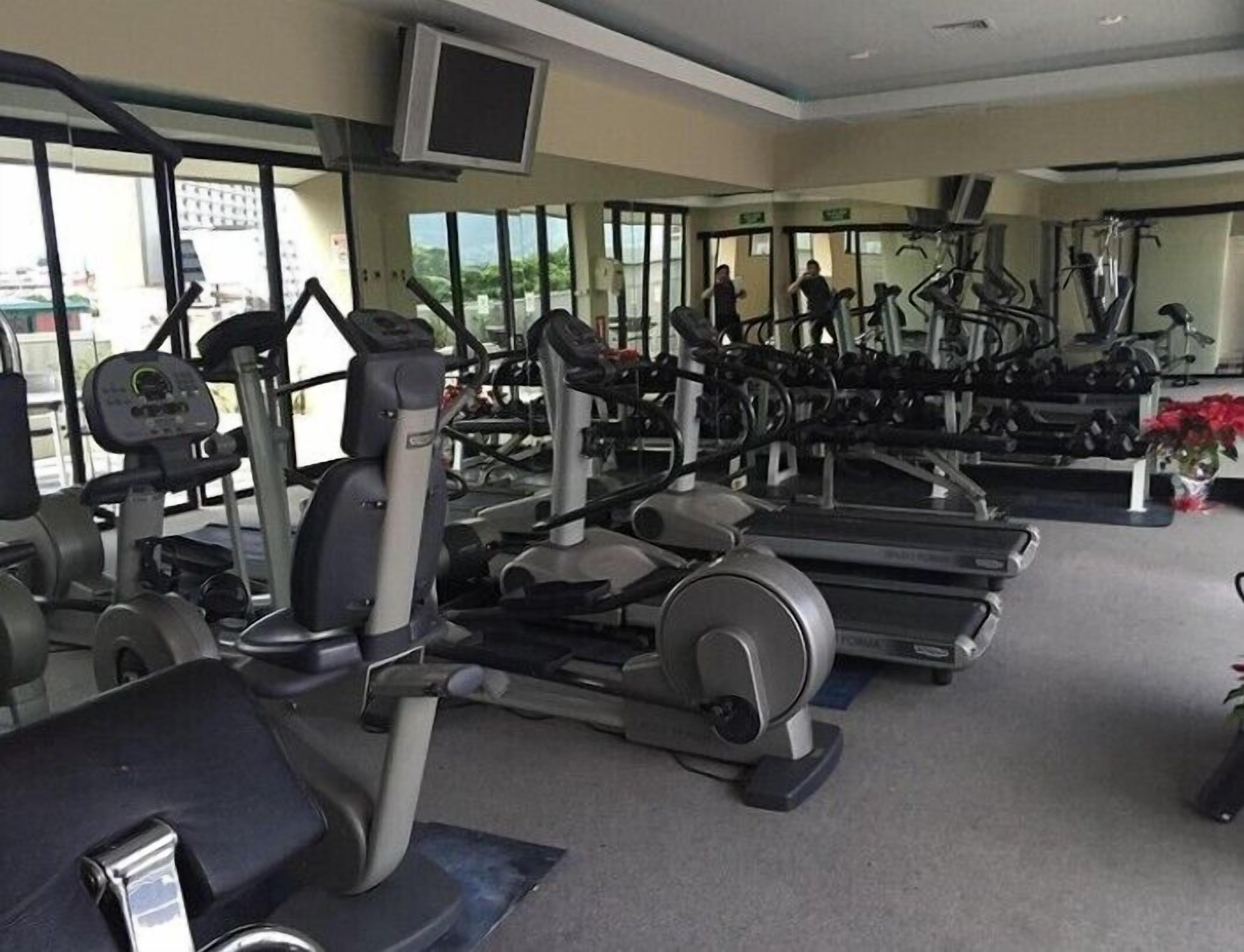 A gym with various exercise equipment, including treadmills and weight machines, and large windows providing natural light.