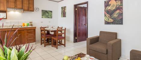 Una sala de estar con un sofá marrón, un comedor con sillas de madera, una cocina con armarios de madera y un frutero sobre una mesa de centro.