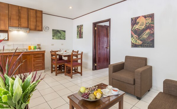 Una sala de estar con un sofá marrón, un comedor con sillas de madera, una cocina con armarios de madera y un frutero sobre una mesa de centro.