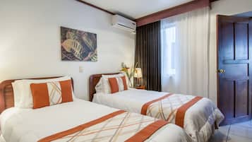 A hotel room with two beds, a painting on the wall, an air conditioning unit, a window with curtains, and a wooden door.