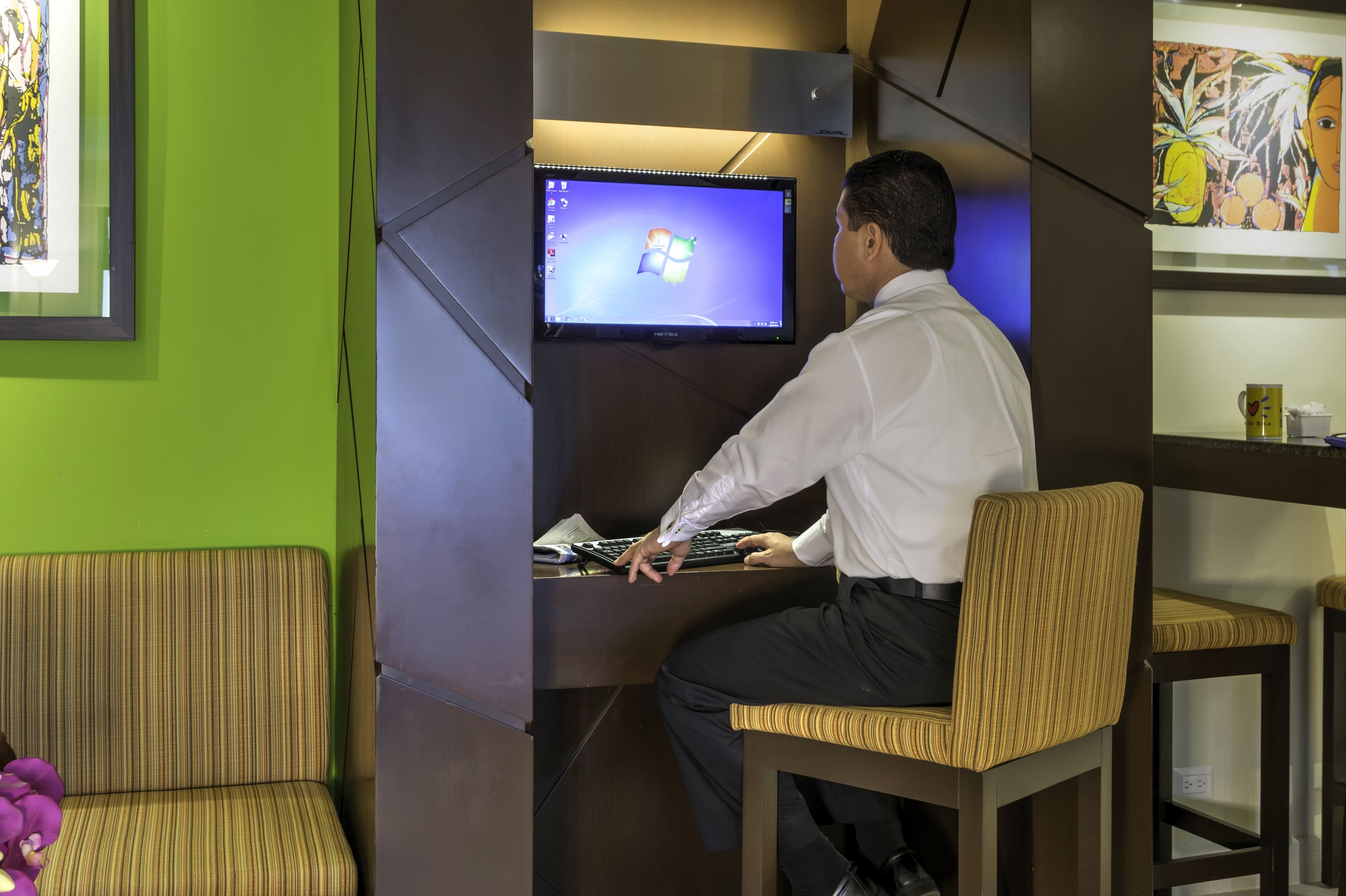 Un hombre sentado en un escritorio con un monitor de computadora mostrando el logotipo de Windows, en una habitación de paredes verdes y una obra de arte enmarcada a la izquierda.