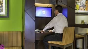 Un hombre sentado en un escritorio con un monitor de computadora mostrando el logotipo de Windows, en una habitación de paredes verdes y una obra de arte enmarcada a la izquierda.