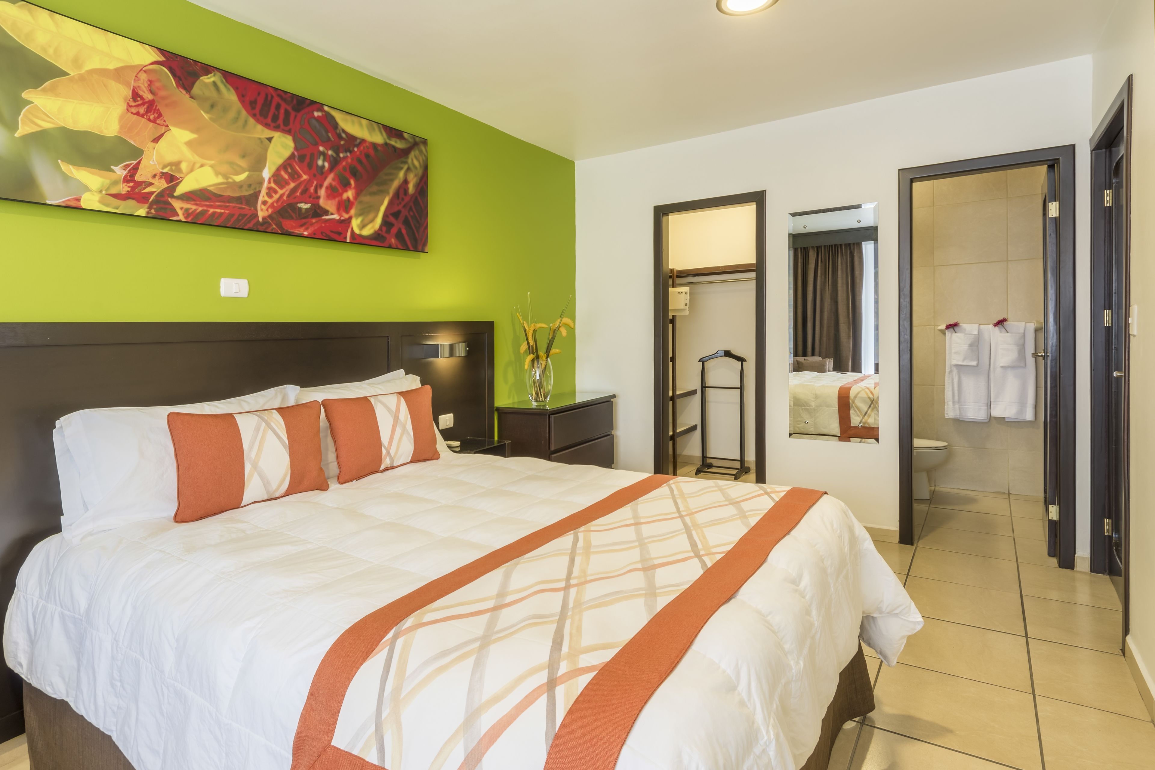 Une chambre d’hôtel avec un grand lit, un mur d’accent de couleur verte, un tableau, une salle de bain avec un miroir et une porte donnant sur un espace douche.