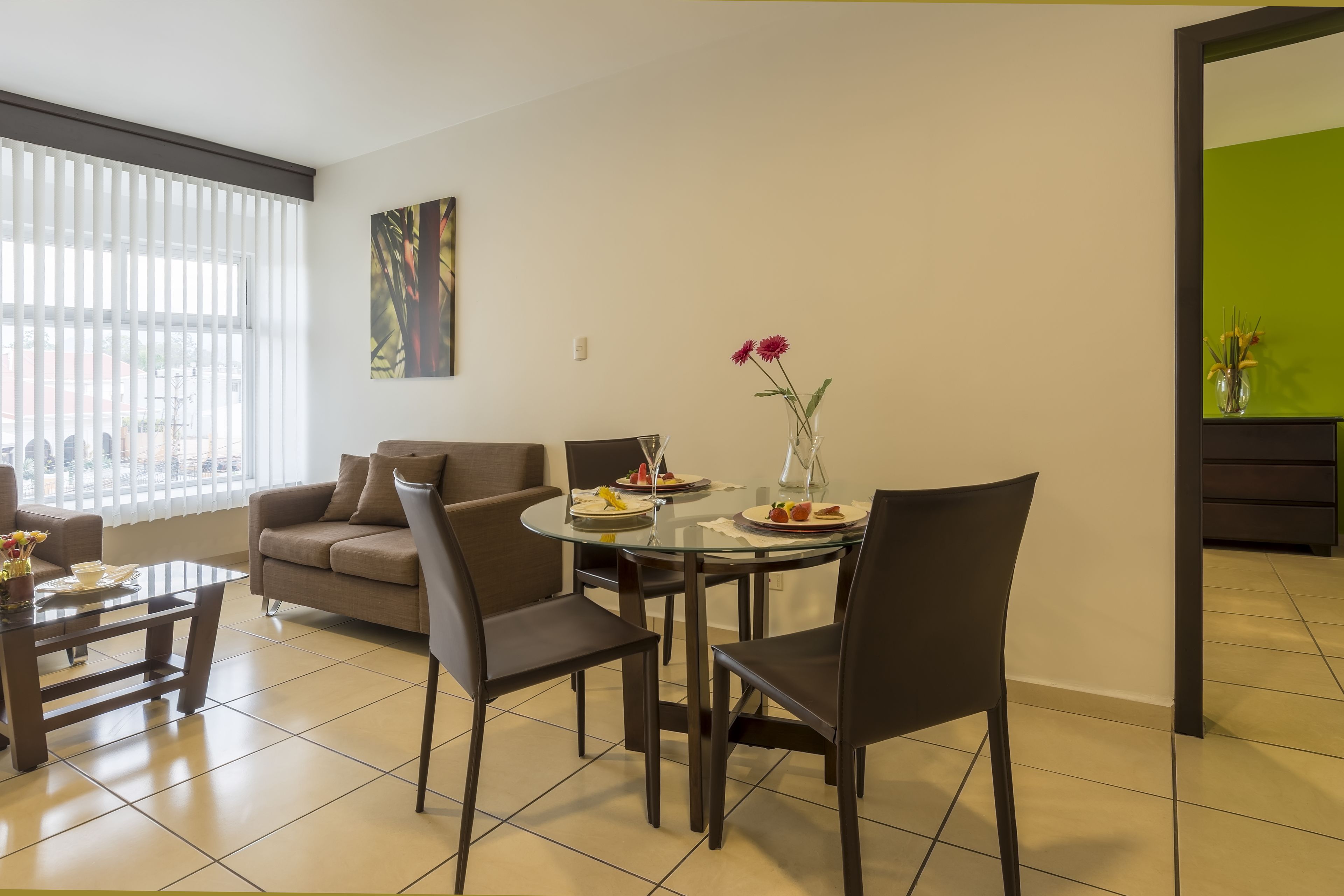 A modern living room with a glass dining table, dark chairs, a sofa, and a vase with flowers.
