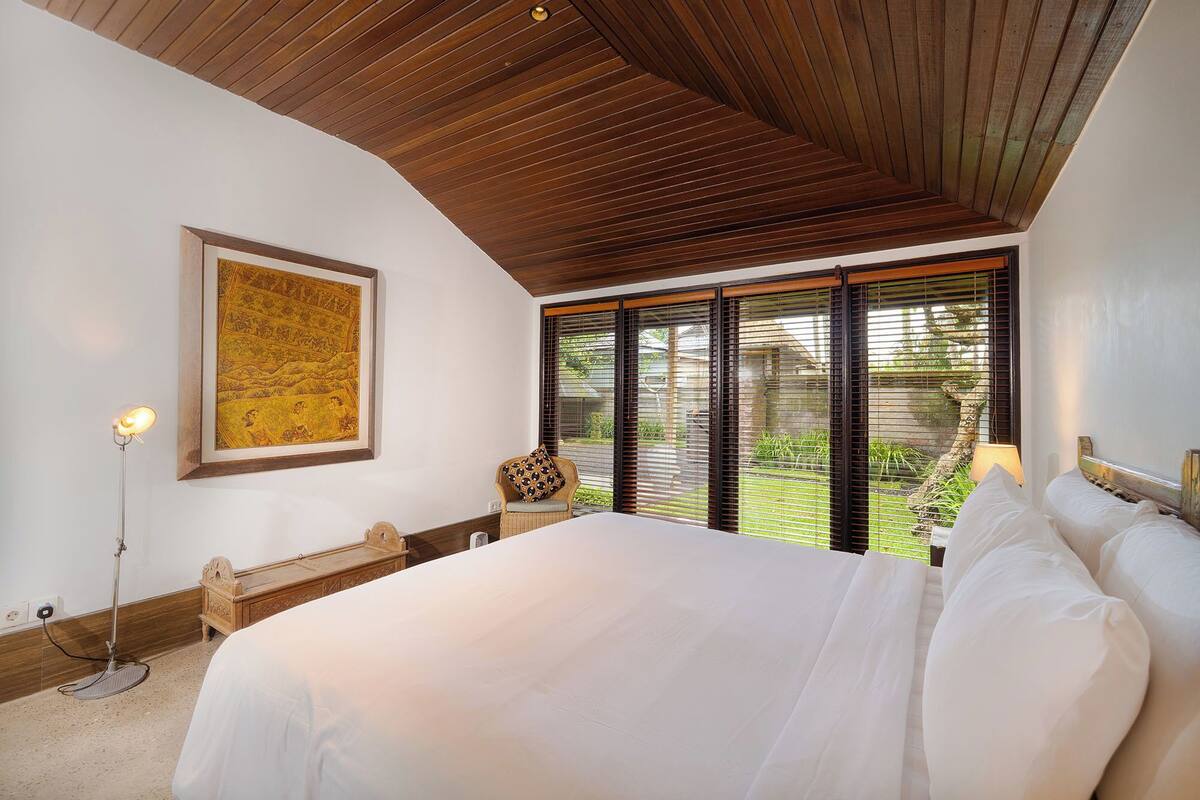 A bedroom with a large bed, a wooden ceiling, a framed artwork, a bedside table, and a view of greenery through the window.