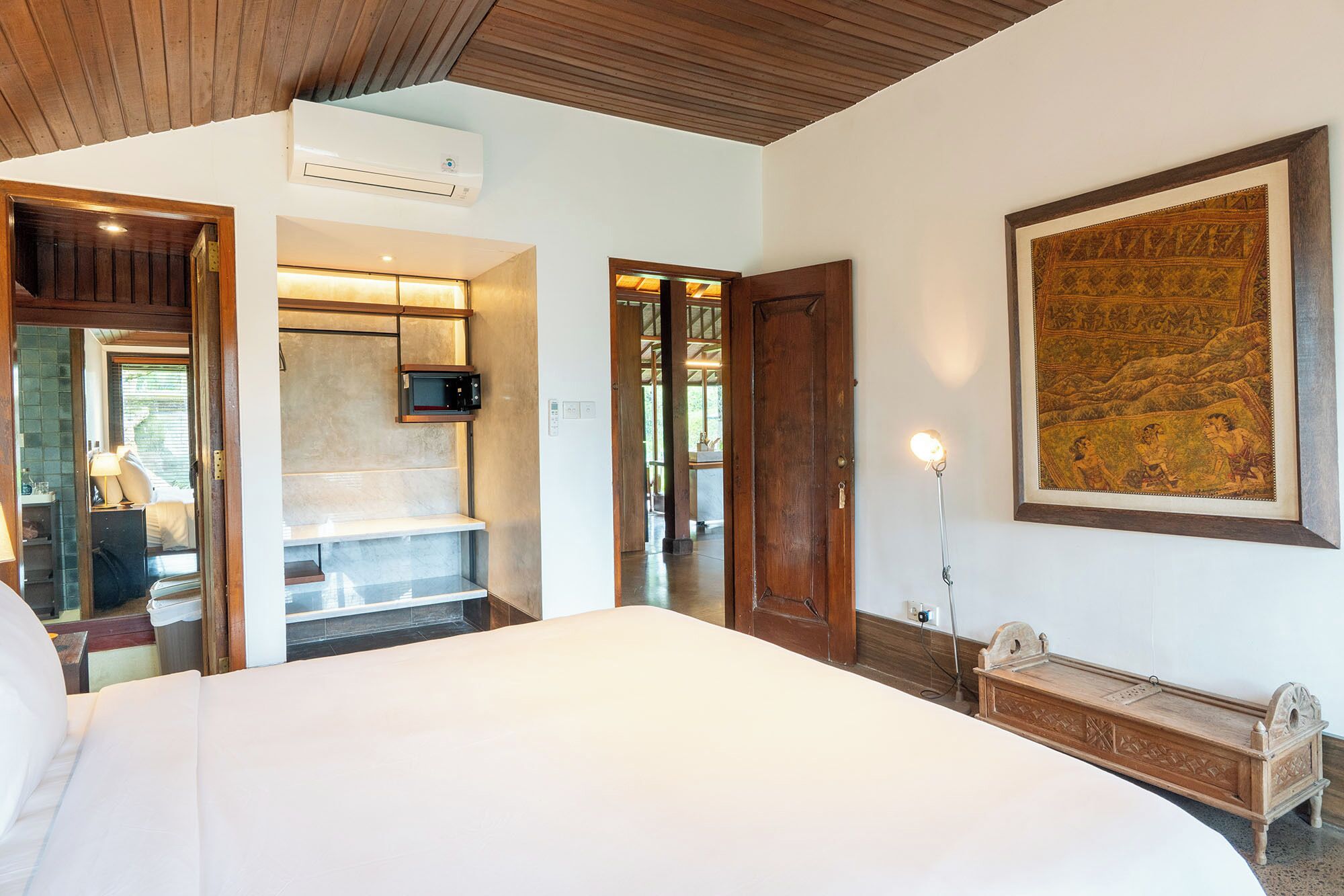 A hotel room with a bed, a wooden ceiling, a mounted TV, a painting, and a bench.