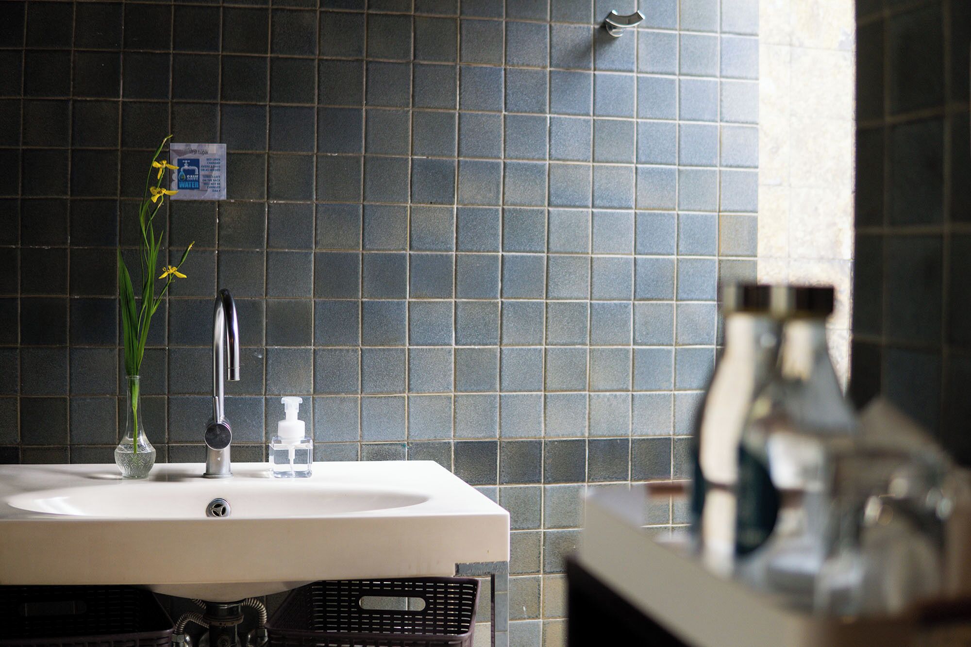 A bathroom with a white sink, a glass vase with a yellow flower, a soap dispenser, and a shower head.
