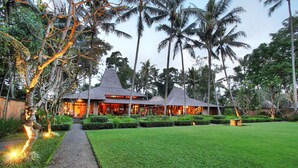 A resort with a well-maintained lawn, palm trees, and a pathway leading to a building with a thatched roof. - Puri Tupai (Ubud)