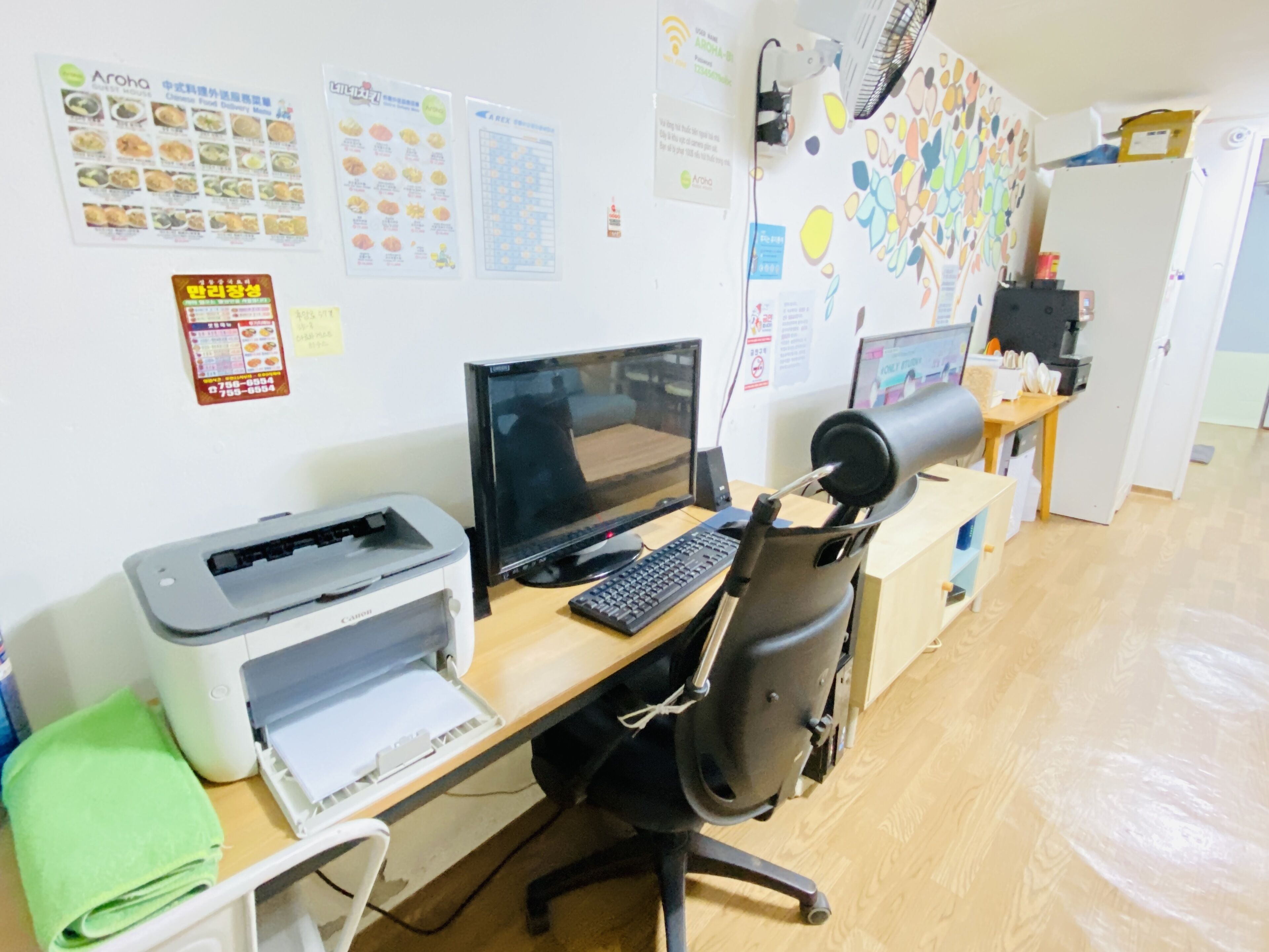 Un espacio de trabajo con computadora de escritorio, impresora y silla de oficina. Hay carteles en la pared y un refrigerador al fondo.