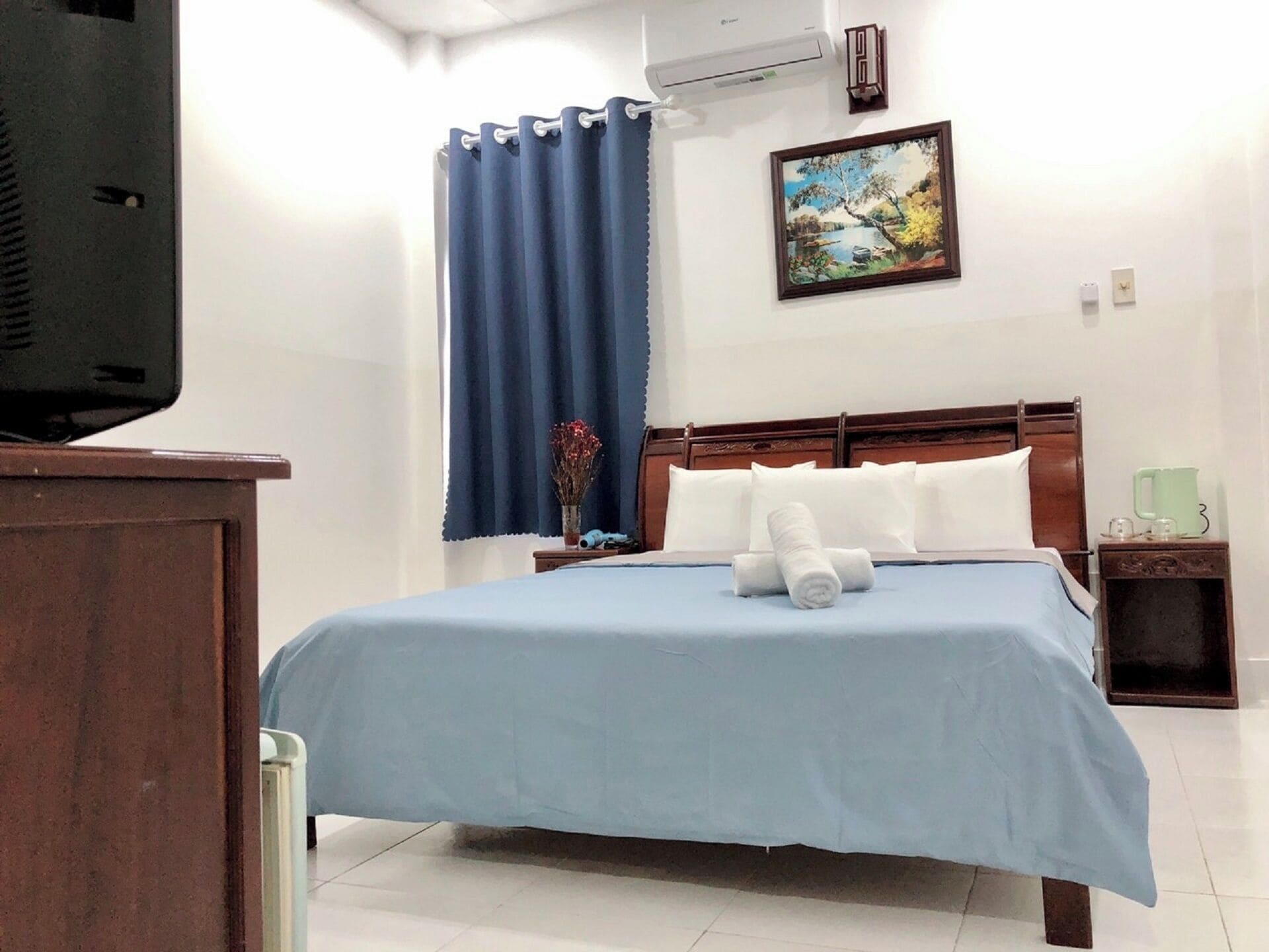 A hotel room with a bed, a nightstand, a TV, a painting, and a window with curtains.