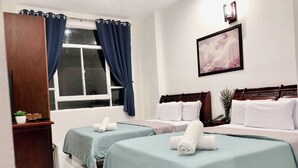 A hotel room with two beds, each with a blue bedspread and white pillows, a wooden headboard, a window with blue curtains, a wall-mounted picture, and a small plant on a wooden shelf. - Saigon Cozy2 Hotel (Ho Chi Minh City)