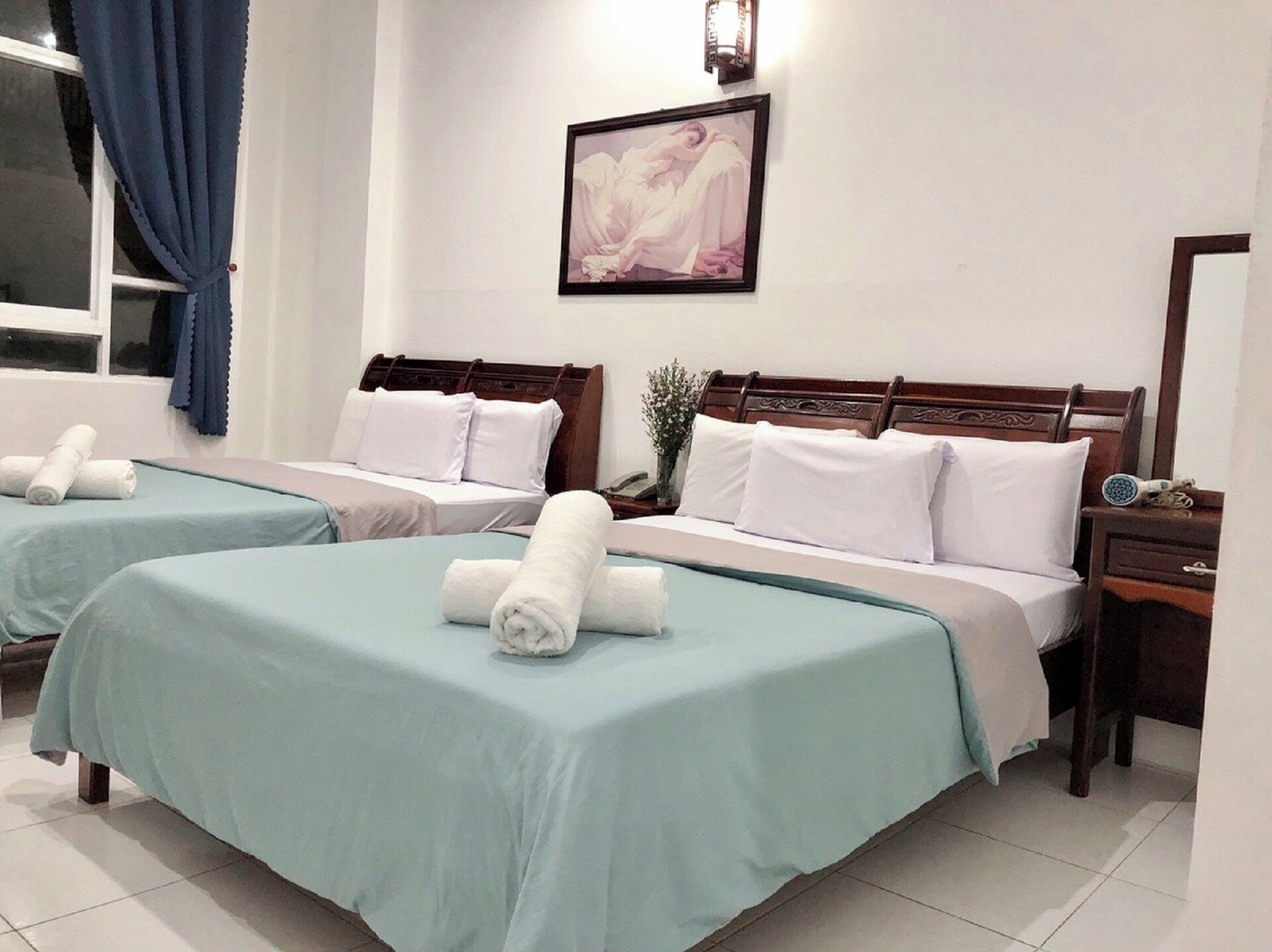 A hotel room with two beds, each with a blue bedspread and white pillows. There is a framed picture on the wall, a window with blue curtains, and a bedside table with a lamp and a hairdryer.
