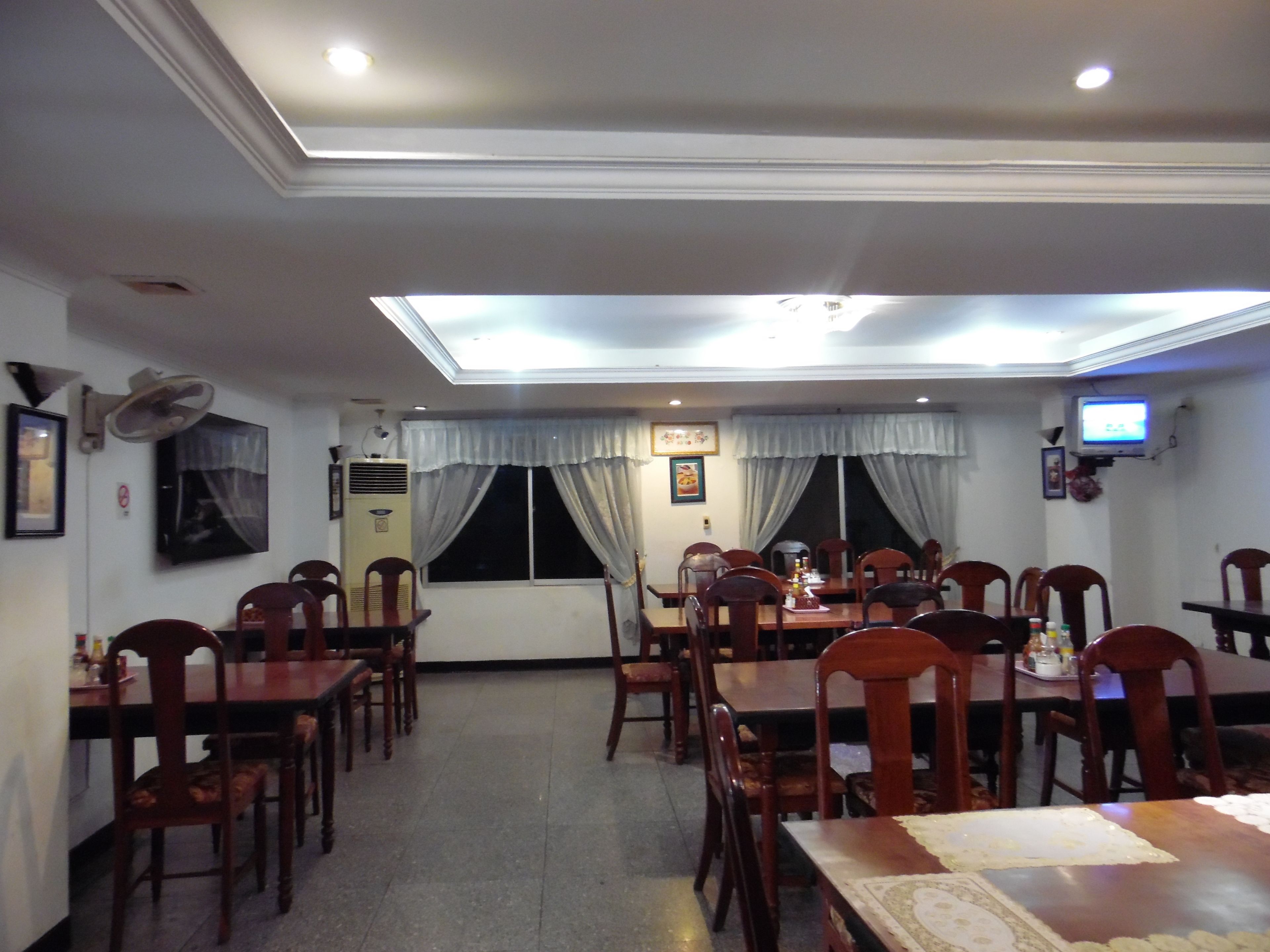 Un comedor con mesas y sillas, un ventilador de techo, un televisor y una ventana con cortinas.