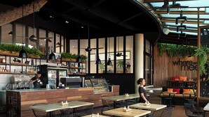 A modern cafe with wooden tables and black wire chairs, a bar area with a person preparing drinks, and a large plant in the background. - Anantara Vacation Club Legian (Legian)