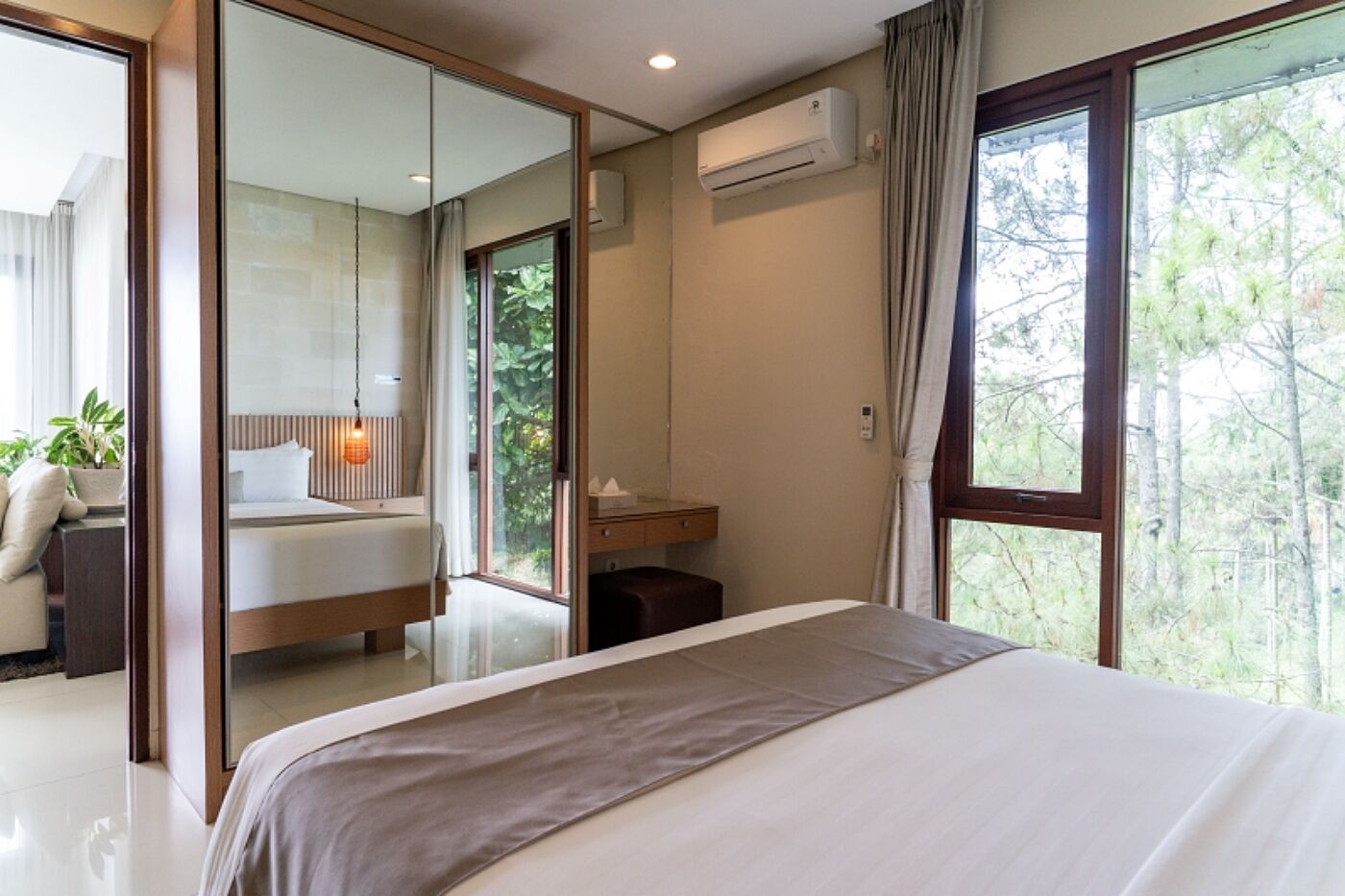 A modern bedroom with a large bed, a mirror, an air conditioning unit, and a view of trees through the window.