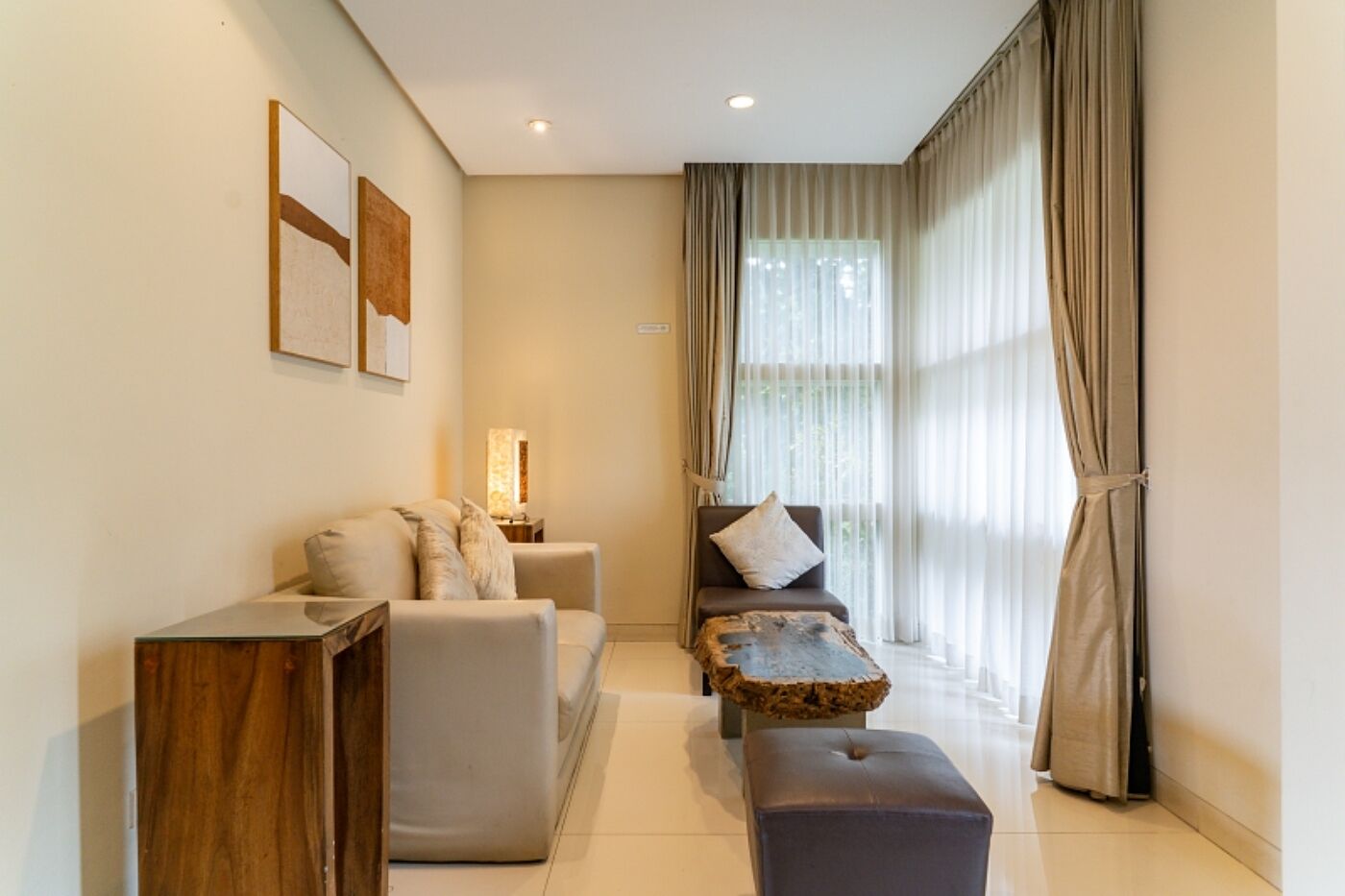 A modern living room with a sofa, ottoman, and a coffee table. There is a lamp on a side table, and curtains on the windows.
