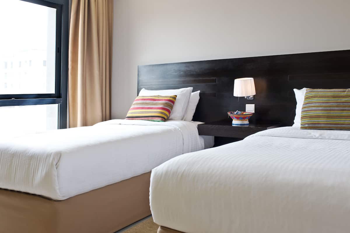 A hotel room with two beds, a nightstand with a lamp, and a window with curtains.