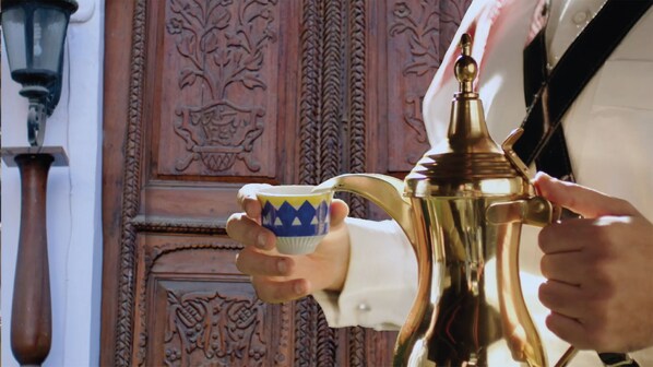 A person holding a coffee pot and a cup, with a decorative wooden door in the background. - Dara Salam (Jeddah)