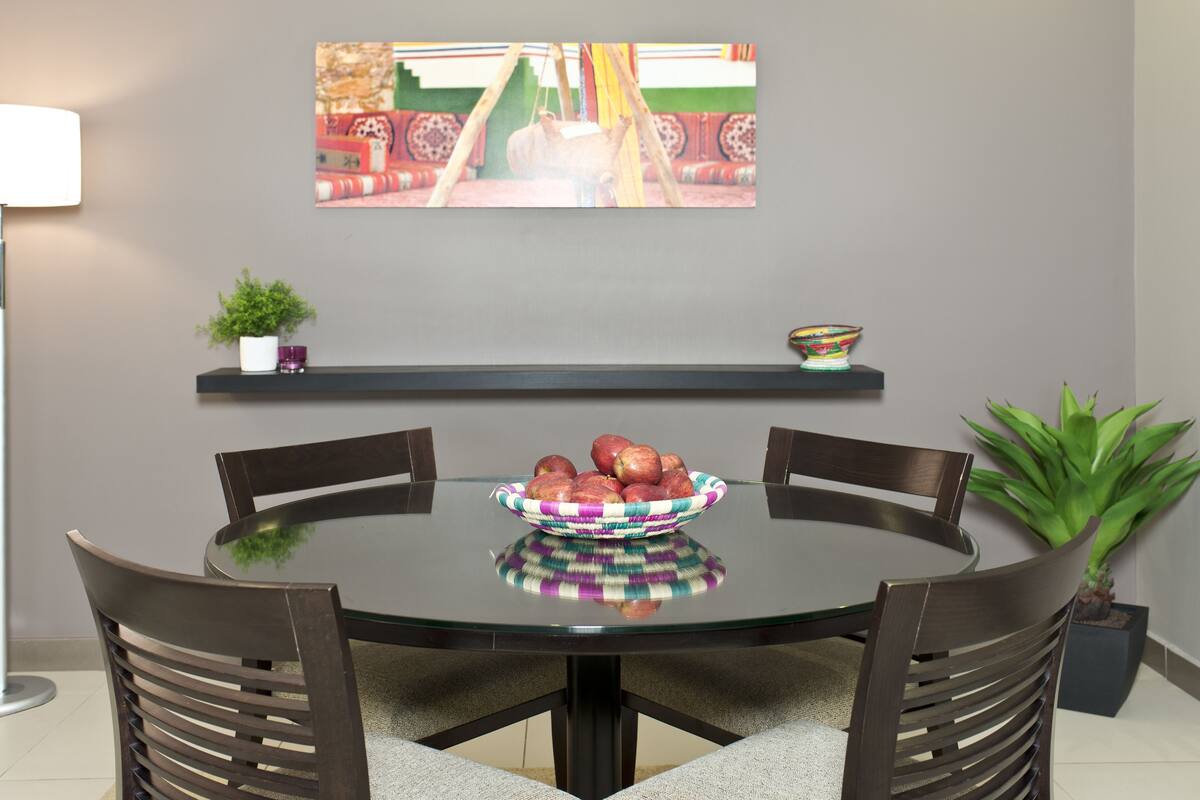 A dining area with a round glass table, four wooden chairs, a bowl of apples, a potted plant, and a shelf with decorative items.