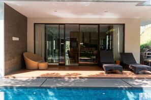 Un espace piscine extérieur moderne doté d’un patio fermé par des baies vitrées, de chaises longues et d’un coin repas visible à travers le verre.