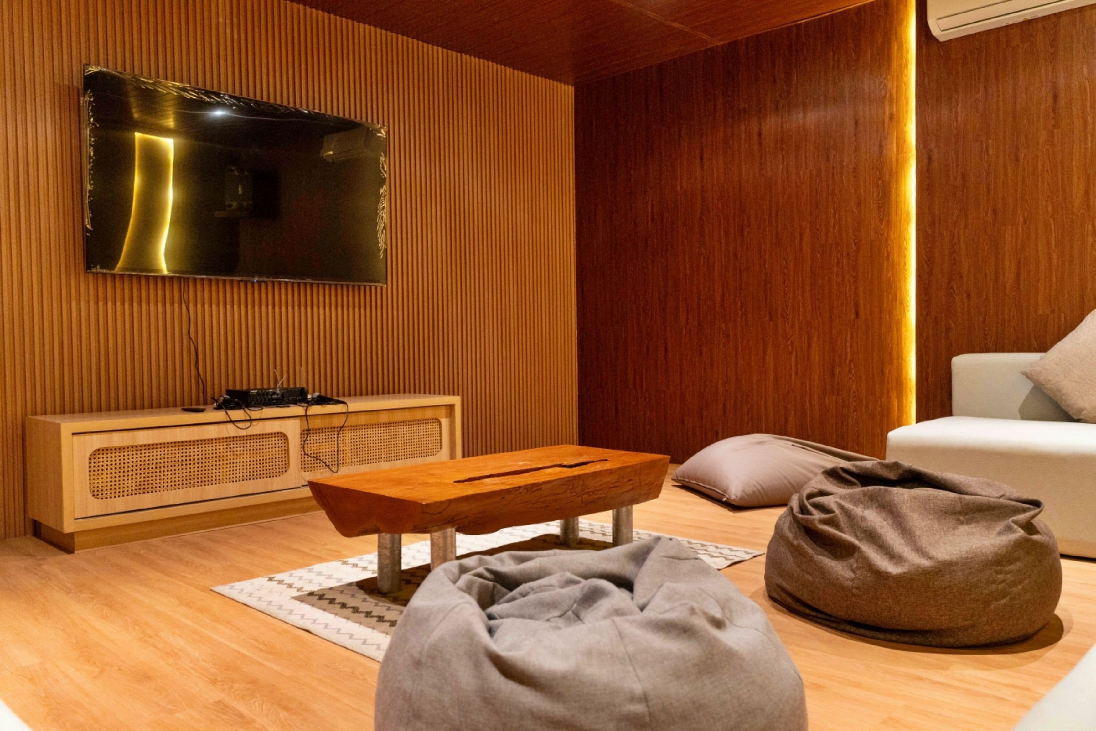Ett modernt vardagsrum med en platt-TV, väggpaneler i trä, ett soffbord i trä, en sittpuff och en vit soffa.