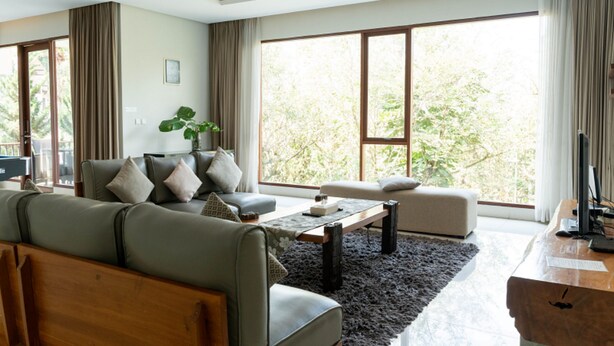 Un salon moderne avec un canapé, un fauteuil et une table basse. De grandes fenêtres sont ornées de rideaux, une plante est présente, et on peut admirer la verdure à l’extérieur.