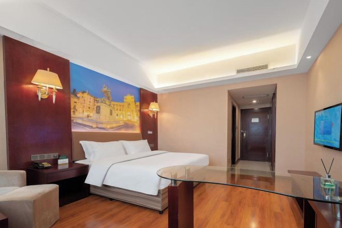 A hotel room with a bed, a glass table, a nightstand, a lamp, a painting on the wall, and a television.