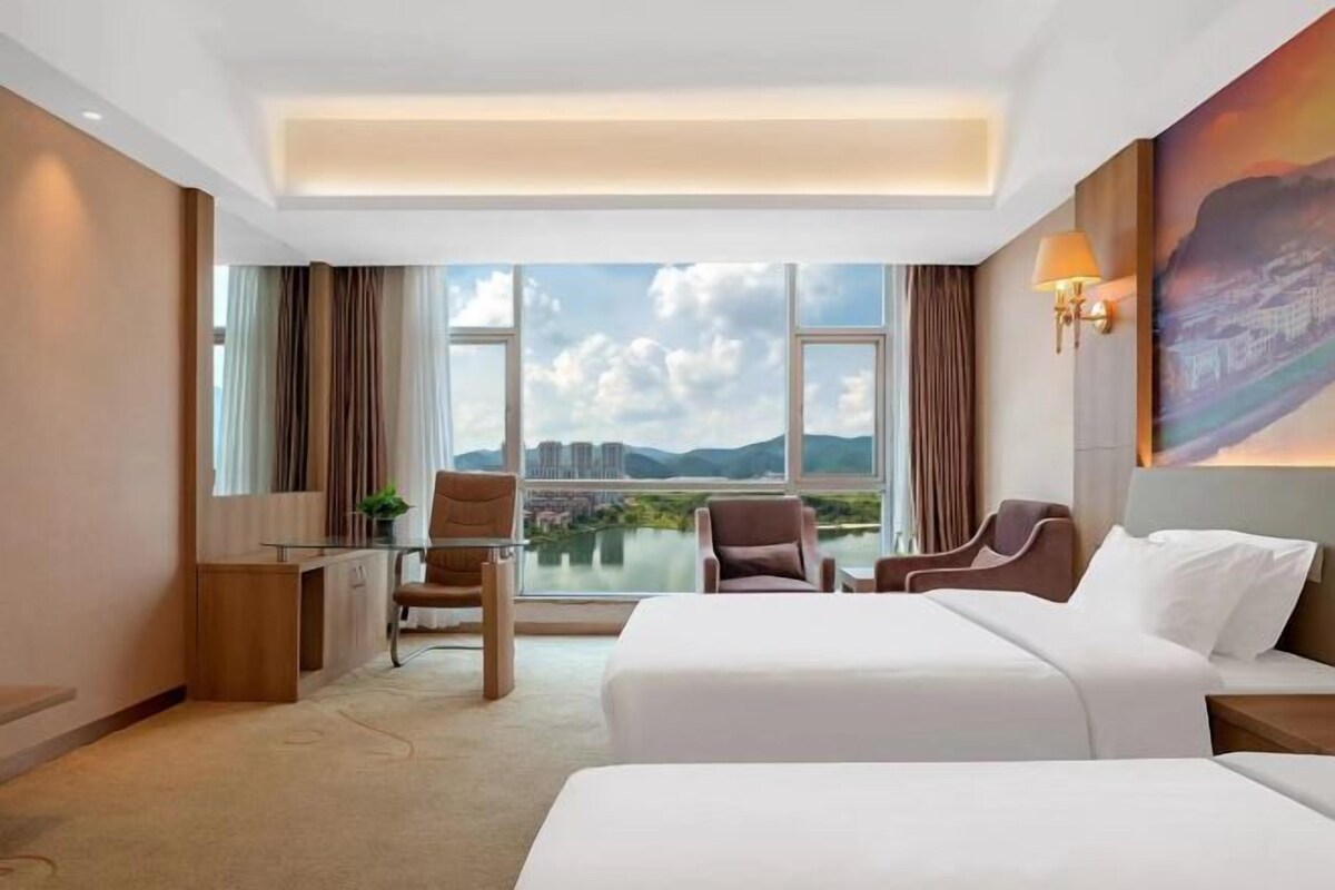 A hotel room with a large window offering a cityscape view, a bed, two chairs, a desk, and a lamp.