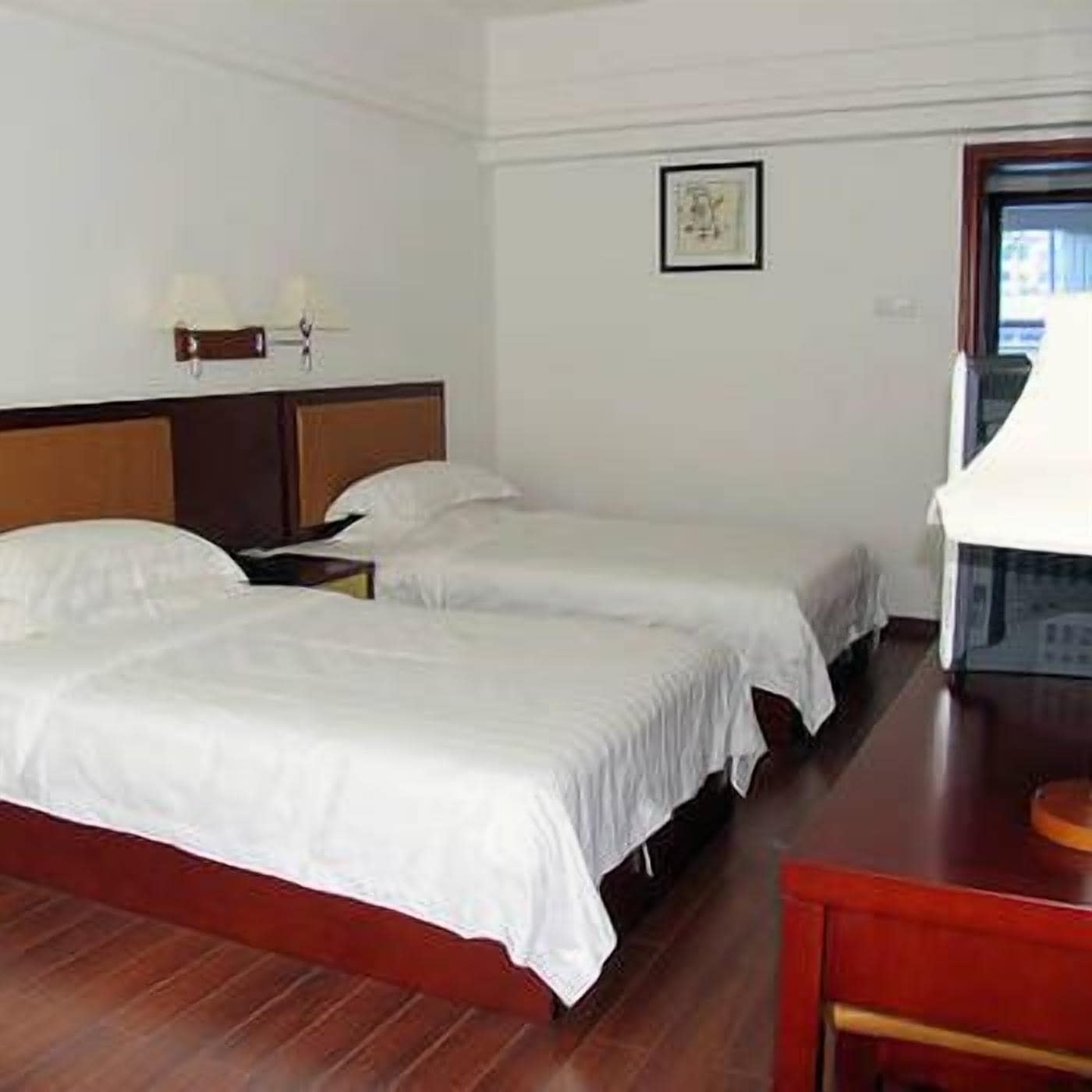 Habitación de hotel con dos camas individuales, cabecera de madera, mesita de noche con lámpara y un cuadro en la pared.