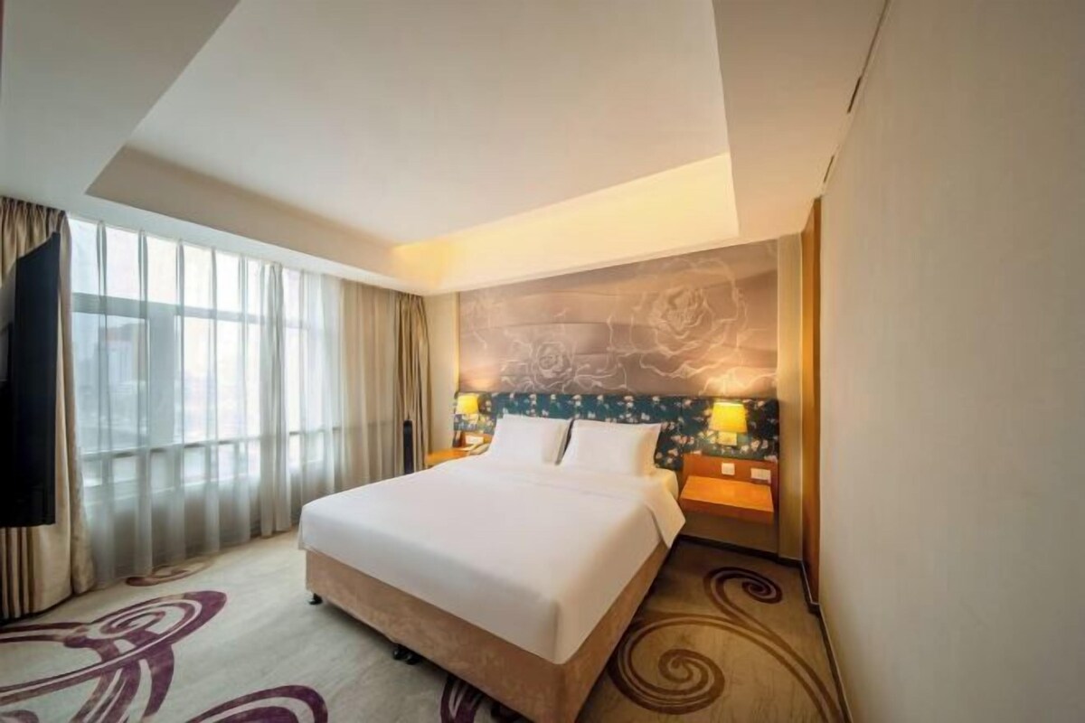Una habitación de hotel con una cama grande, un cabecero con diseño floral, una mesita de noche con lámpara y vistas a la ciudad a través de amplios ventanales.