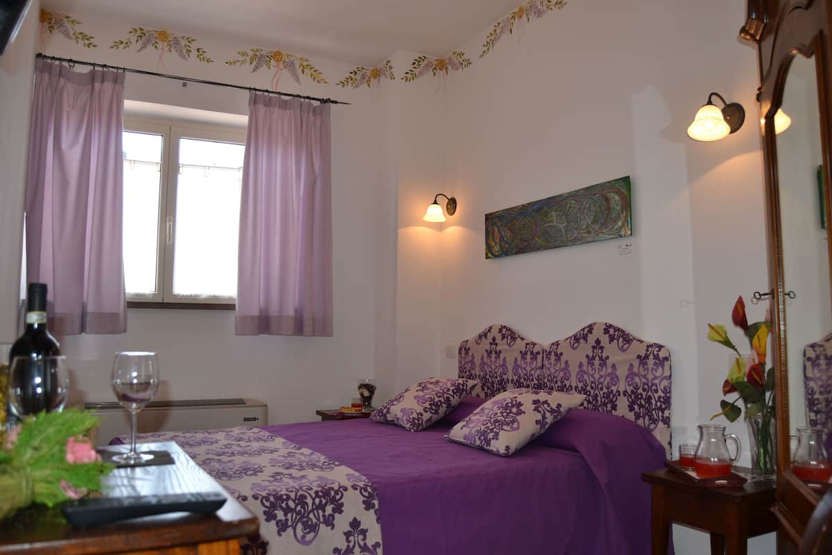 A bedroom with a bed, curtains, a painting, a bedside table with a glass, and a mirror.